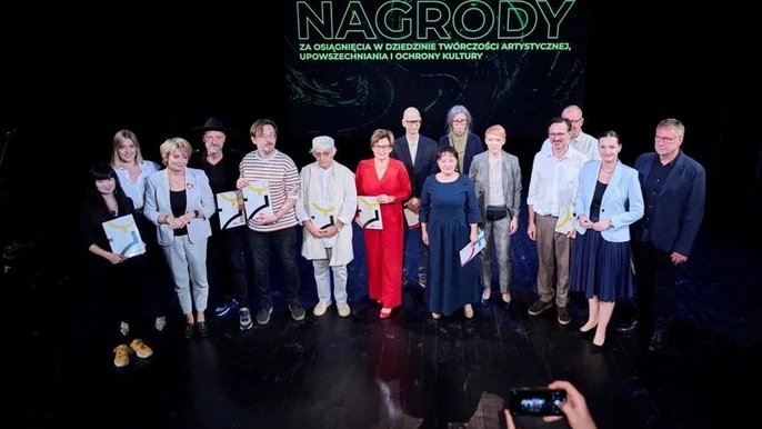  - Na scenie teatralnej stoi grupa ludzi, którzy w rękach trzymają dyplomy. Są to laureaci nagród w 2023 roku. W tle napis: Nagrody za osiągnięcia w dziedzinie twórczości artystycznej, upowszechnia i ochrony kultury.